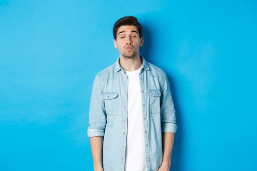 Handsome man waiting for kiss, pucker lips and close eyes while standing against blue background.