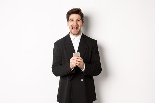 Portrait of happy good-looking man, wearing black suit, laughing from happiness and using mobile phone, standing over white background.