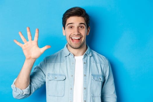 Close-up of handsome man smiling, showing fingers number five, standing over blue background.