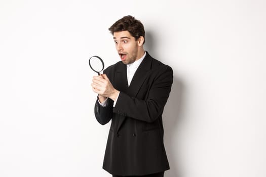 Handsome businessman in black suit, holding magnifying glass and smiling, found something, standing against white background.
