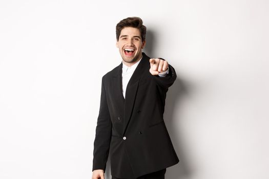 Concept of new year party, celebration and lifestyle. Portrait of handsome stylish man in black suit, smiling and pointing finger at camera, standing over white background.