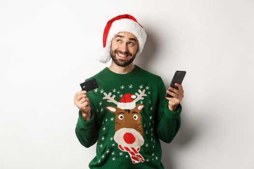 Online shopping and winter holidays concept. Smiling man thinking and holding credit card with mobile phone, wearing Santa hat, white background.