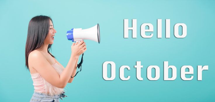 Hello October, Asian happy portrait beautiful young woman standing smile her holding her Shouting through a megaphone message to say "Hello October" isolated on blue background
