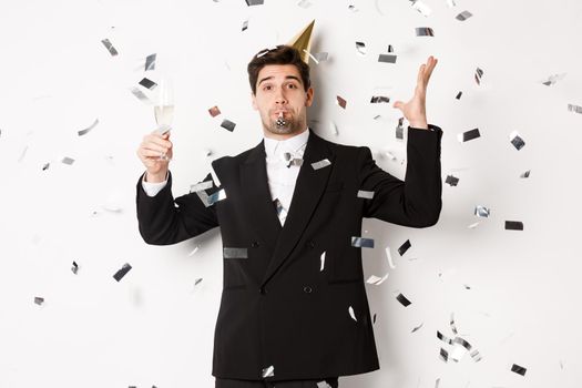 Handsome party guy in black suit having fun, celebrating new year, blowing whistle and drinking champagne while confetti falling, wishing happy holidays, standing against white background.