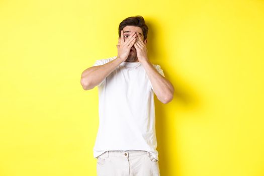 Embarrassed guy shut eyes but peeking through fingers at something awkward, standing over yellow background.