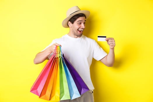 Excited man going shopping on vacation, looking satisfied at credit card, holding paper bags, standing over yellow background.