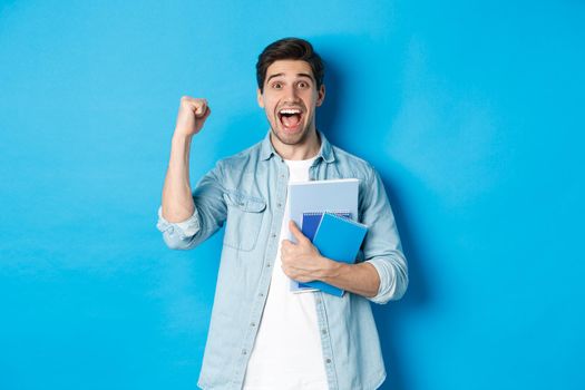 Cheerful guy holding notebooks and celebrating, making fist pump and shouting yes with excitement, standing over blue background.