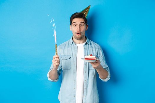 Happy young man celebrating birthday in party hat, holding b-day cake and smiling, standing over blue background.