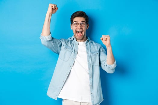 Excited handsome man triumphing, raising hands up and shouting for joy, celebrating victory, standing against blue background.