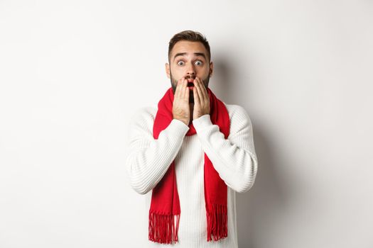Christmas holidays and celebration concept. Handsome bearded guy in red scarf and sweater looking shocked, impressed with advertisement, white background.