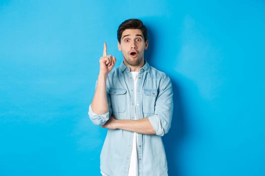 Image of handsome adult man having an idea, raising finger and smiling excited, found solution, standing against blue background.