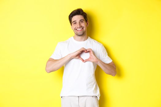 Happy romantic man showing heart sign, smiling and express love, standing over yellow background. Copy space