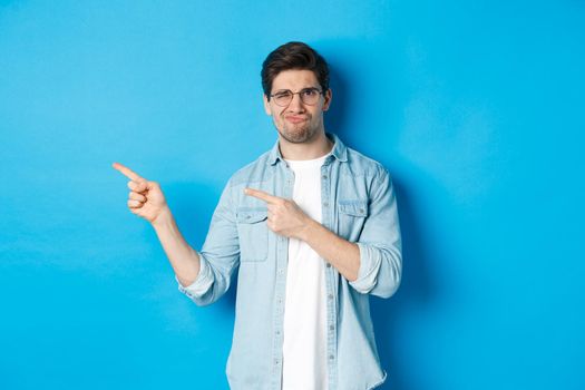 Displeased and skeptical male model in glasses, pointing fingers left at something bad, showing awful advertisement, standing over blue background.