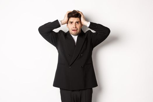 Frustrated and worried businessman in black suit, panicking as looking at trouble, holding hands on head alarmed, standing against white background.
