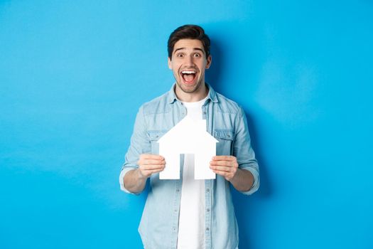 Insurance, mortgage and real estate concept. Happy man holding house model and smiling excited, buying property or renting apartment, standing against blue background.