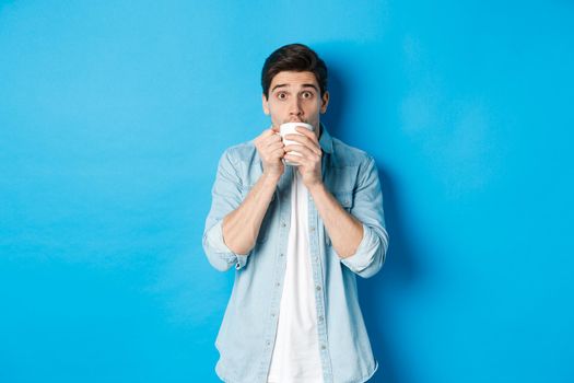 Man looking excited and sipping tea or coffee from white mug, standing over blue background.