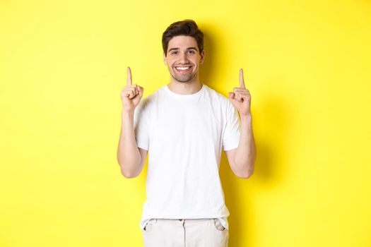 Handsome guy in white t-shirt pointing fingers up, showing shopping offers, standing over yellow background.
