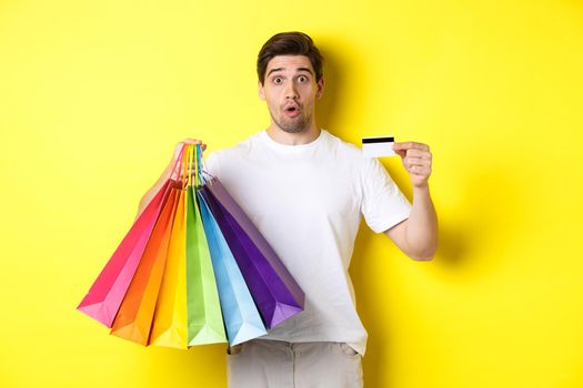 Excited man shopping on black friday, holding paper bags and credit card, standing against yellow background.