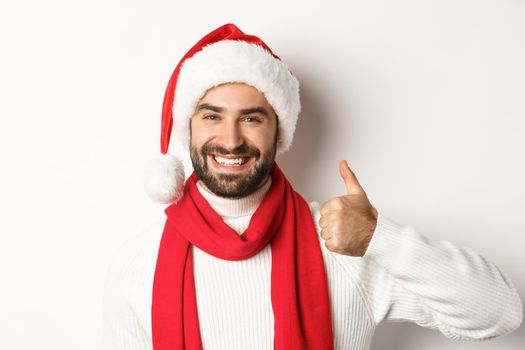 New Year party and winter holidays concept. Close-up of happy man in Santa hat showing thumbs up in approval, like and agree, standing over white background. Copy space