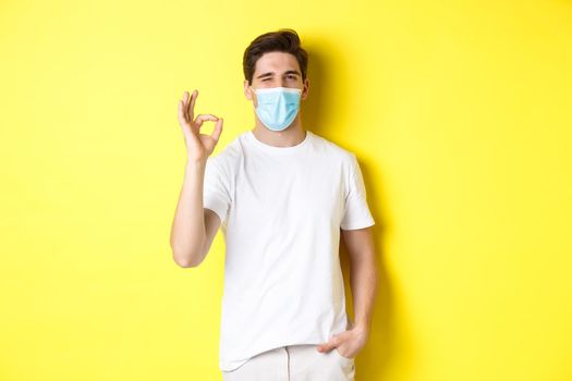 Concept of coronavirus, pandemic and social distancing. Confident young man in medical mask showing okay sign and winking, yellow background.