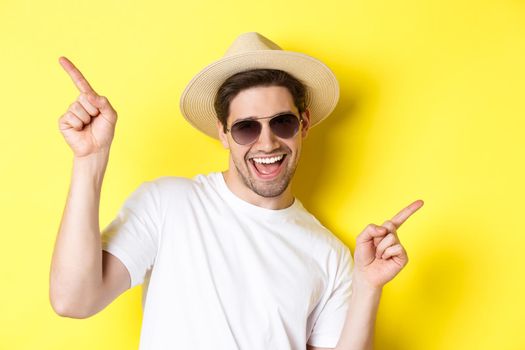 Concept of tourism and vacation. Close-up of man enjoying holidays on trip, dancing and pointing fingers sideways, wearing sunglasses with straw hat.
