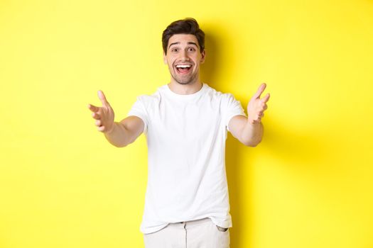 Excited handsome guy stretching hands forward, reaching for hug, receiving gift, standing over yellow background.