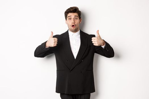Concept of new year party, celebration and lifestyle. Portrait of excited handsome businessman in formal suit, showing thumbs-up in approval, recommending shop, standing over white background.