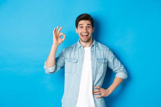 Smiling happy man showing ok sign and looking pleased, approving something good, standing against blue background.