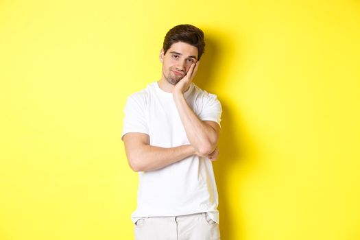 Bored and unamused man looking with indifference, standing in white clothes over yellow background.