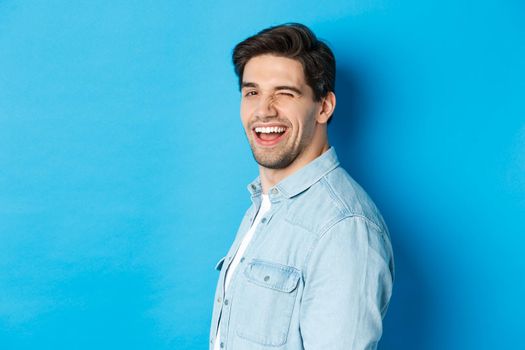 Happy and confident man turn head at camera, winking and smiling, standing over blue background.