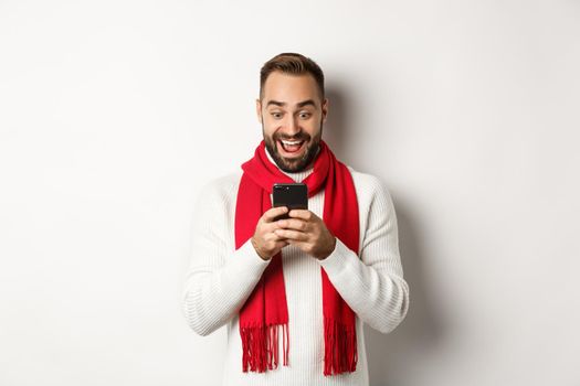 Man reading messaging on mobile phone and looking happy, standing in winter sweater and red scarf, white background.