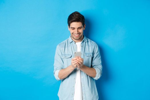 Image of handsome young man using mobile phone, texting on phone and looking pleased, standing over blue background.