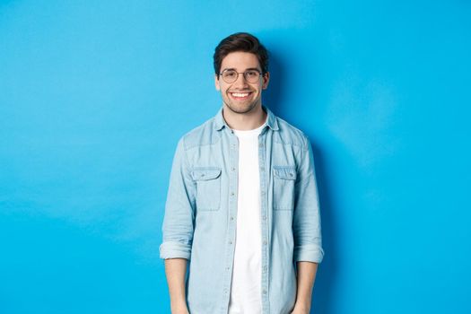 Young modern man in glasses and casual outfit standing against blue background, smiling happy at camera.