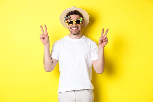 Concept of tourism and lifestyle. Happy man enjoying trip, wearing summer hat and sunglasses, posing with peace signs for photo, yellow background.