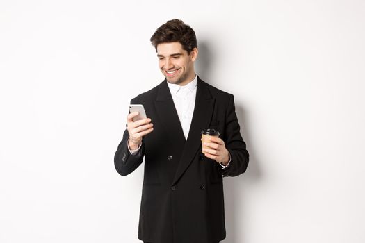 Portrait of handsome, confident businessman in black suit, drinking coffee and using mobile phone, smiling pleased, standing over white background.