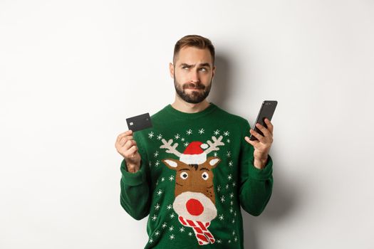 New Year, online shopping and christmas concept. Thoughtful man using mobile phone and credit card, thinking about gifts, standing over white background.