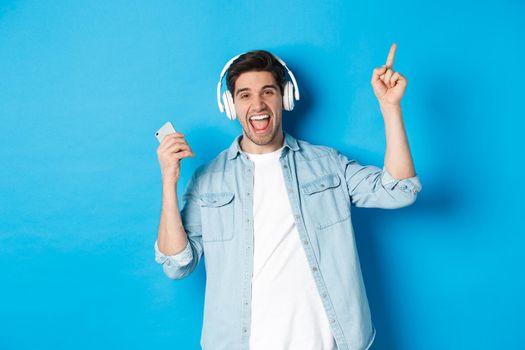 Joyful handsome man dancing with smartphone, listening music in headphones and pointing finger up, standing over blue background.