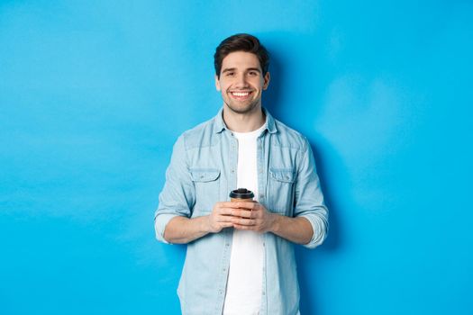 Young happy man drinking coffee from cafe takeaway, smiling pleased, standing against blue background.