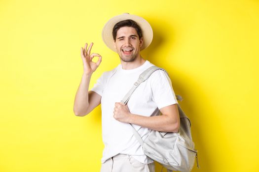 Tourism, travelling and holidays concept. Happy tourist going on vacation, holding backpack and showing okay sign smiling satisfied, standing against yellow background.