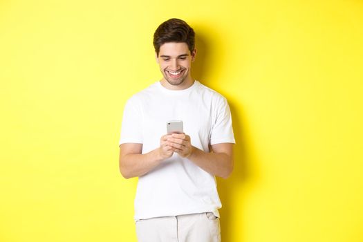 Young man reading text message on smartphone, looking at mobile phone screen and smiling, standing in white t-shirt against yellow background.