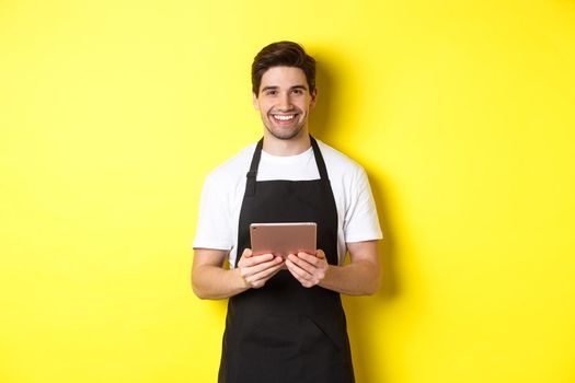 Waiter in black apron taking orders, holding digital tablet and smiling friendly, standing over yellow background.