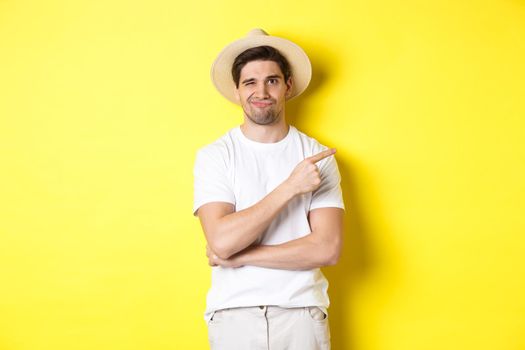 Skeptical male tourist complaining, pointing finger right at something bad or lame, standing unamused against yellow background.