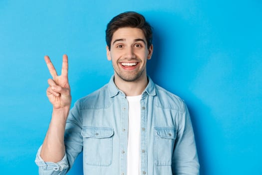 Close-up of handsome man smiling, showing fingers number two, standing over blue background.