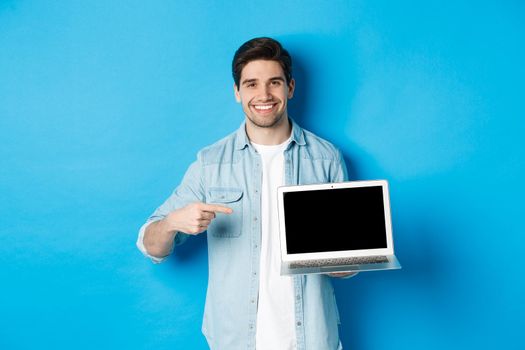 Handsome young man pointing finger at screen of computer, smiling pleased, showing promo in internet or website, standing over blue background.