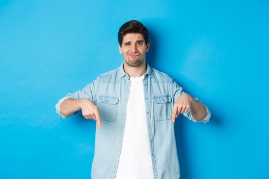 Disappointed guy in casual outfit, pointing fingers down with doubtful grimace, standing against blue background.