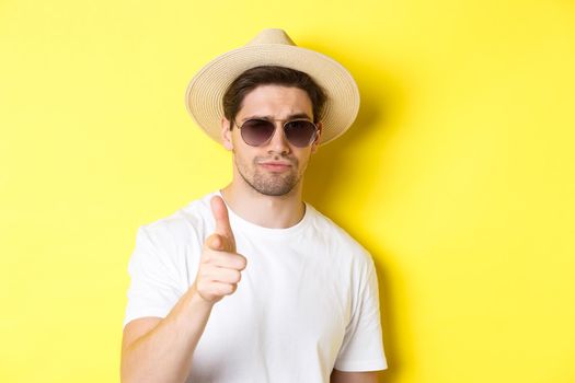 Concept of tourism and vacation. Close-up of cool guy in summer hat and sunglasses pointing finger at camera, standing over yellow background.