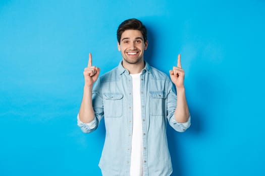 Portrait of handsome bearded man in casual outfit, smiling happy and pointing fingers up at copy space, standing over blue background.