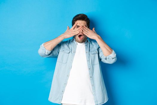 Shocked man covering eyes and peeking through fingers, stare at something embarrassing, standing against blue background.