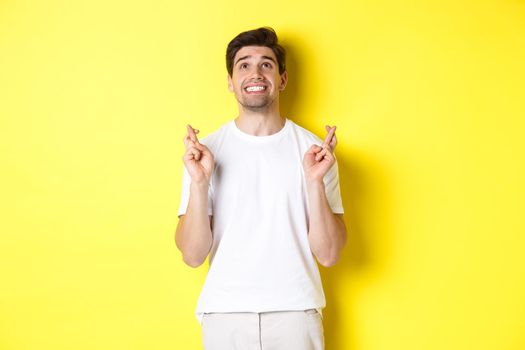 Nervous and hopeful man praying to god, making wish with fingers crossed, panicking and standing over yellow background.