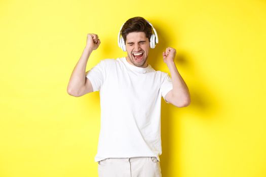 Happy man dancing and listening music in white headphones, standing over yellow background. Copy space